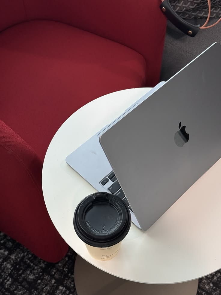 A laptop on a white table next to a coffee and a red armchair